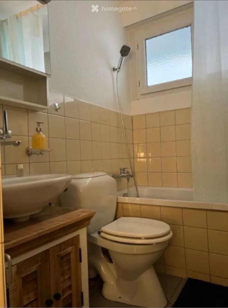 Bathroom with beige tiles, a sink, toilet, bathtub, and a showerhead. Features a wooden cabinet and a mirror above the sink.