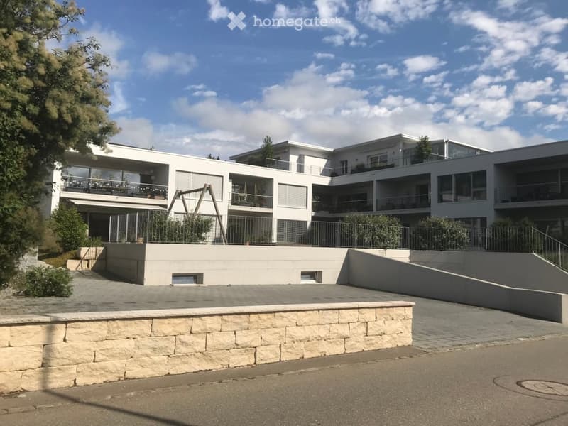 Modern multi-story residential building with white facade, large windows, balconies, and raised ground floor.