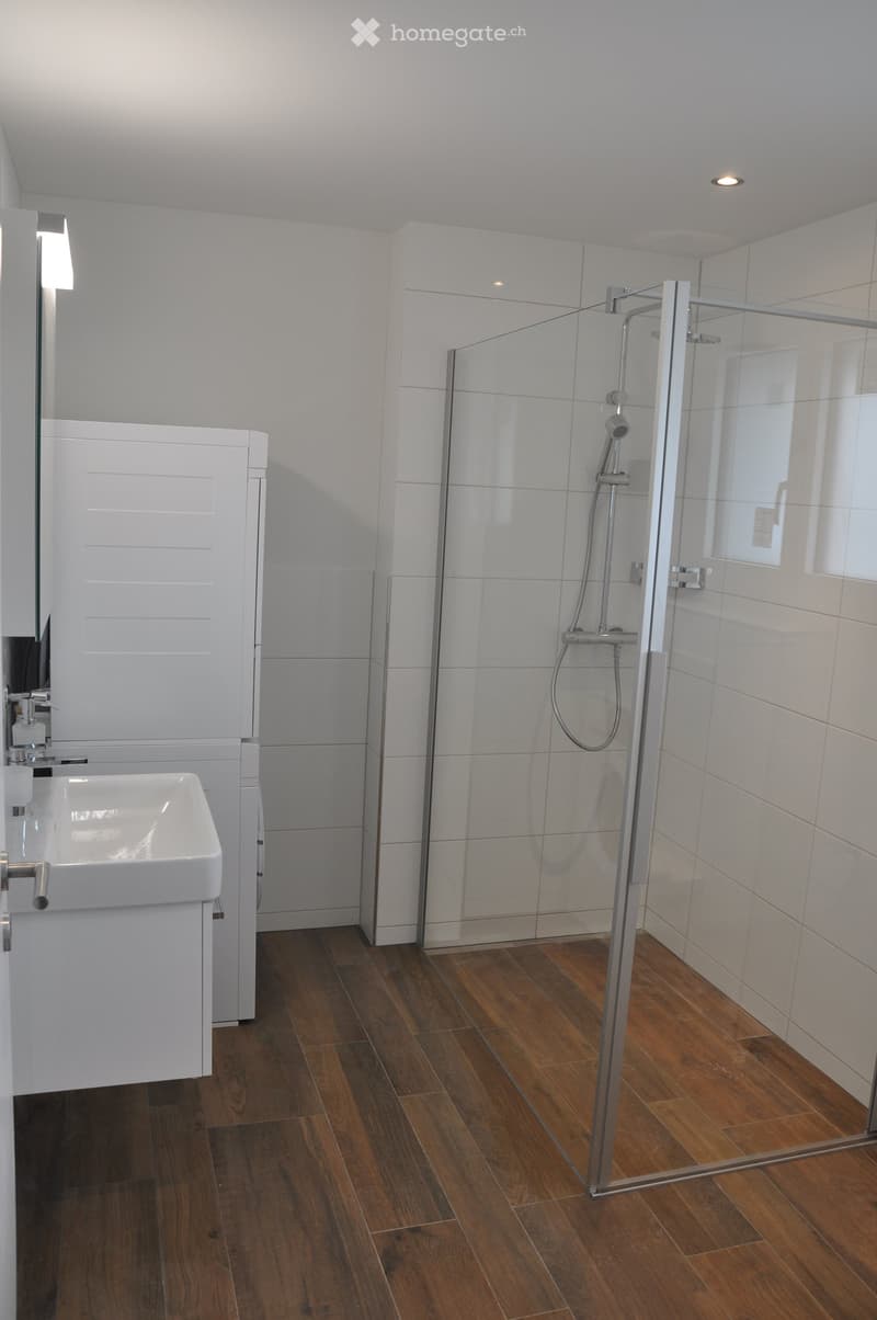 Modern bathroom with wooden flooring, white tiled walls, a glass-enclosed shower, a white sink, and a mirrored cabinet.