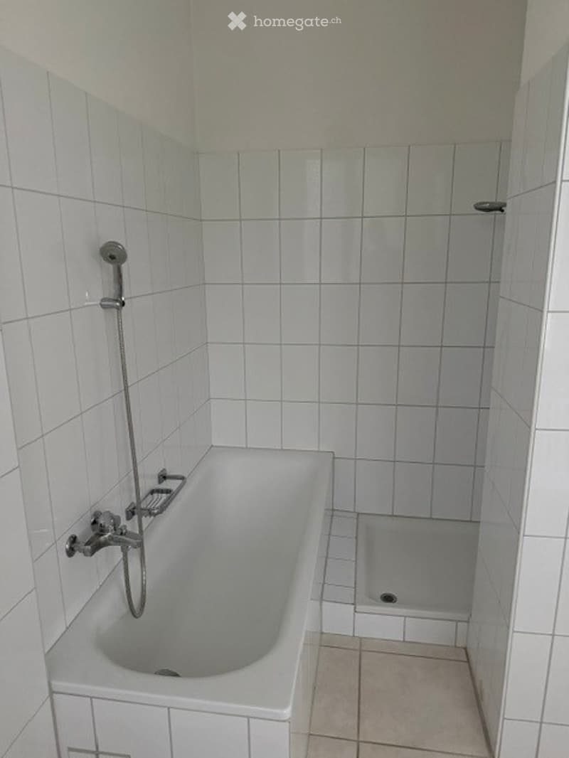 A simple bathroom with white tiled walls and floor, featuring a bathtub with a showerhead and a small shower rack.