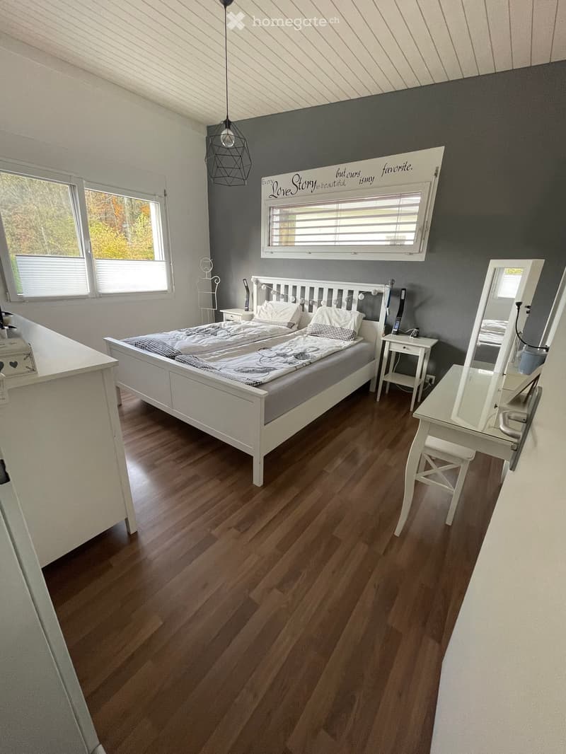Cozy bedroom with wooden flooring, a white bed with pillows and blankets, a nightstand, a mirror, and a small table with a chair.