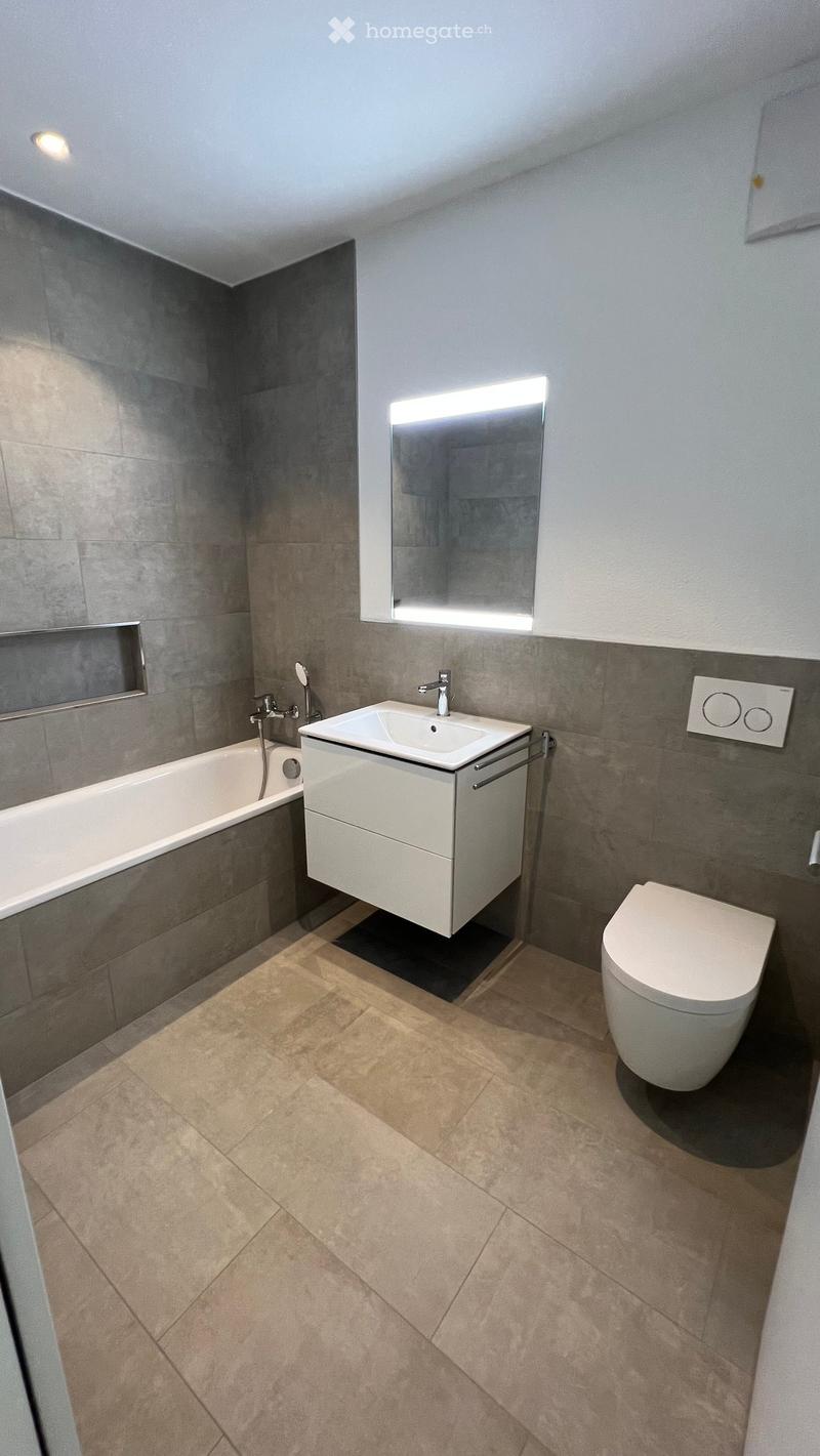 Modern bathroom with a bathtub, sink, toilet, and large mirror with built-in lighting. Grey tiled walls and beige tiled floor.
