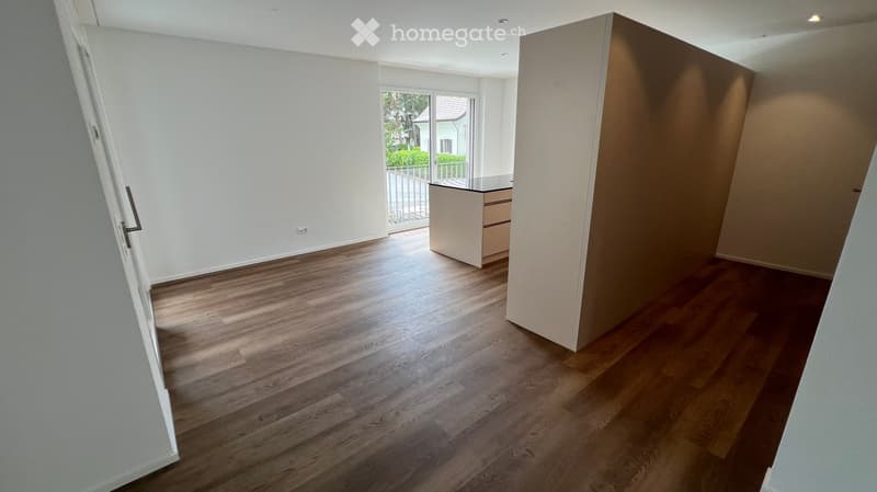 Empty room with wooden flooring, white walls, and a small built-in cabinet. The room is well-lit with natural light coming from a large window with a view of the garden.