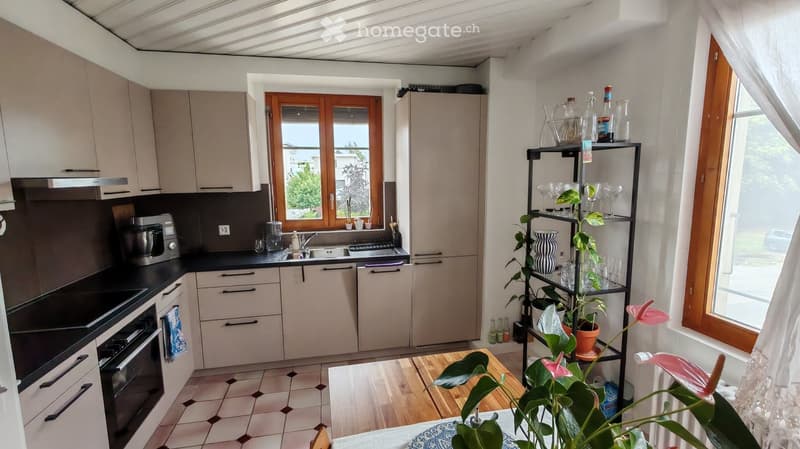 Modern kitchen with light-colored cabinets, black countertops, and stainless steel appliances. Features a window with wooden frame, a sink, and a shelf with glassware and plants.