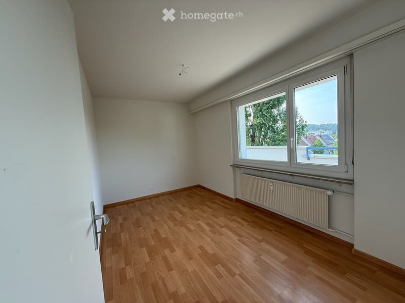 Empty room with wooden flooring, white walls, large window with view, radiator, and door handle visible.