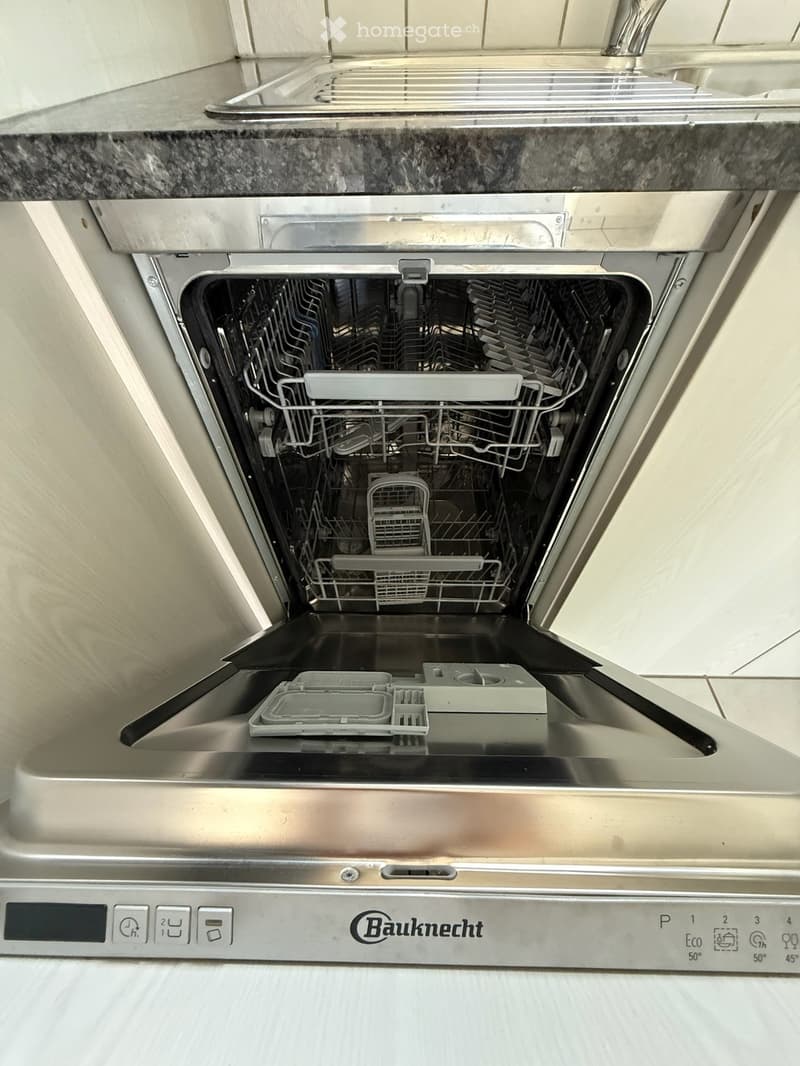 Open stainless steel dishwasher with multiple racks and compartments, installed under a granite countertop.