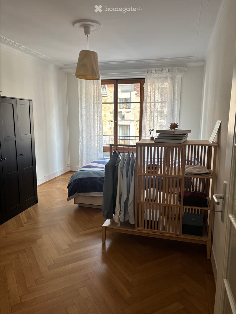 A modern bedroom with wooden flooring, a large bed with blue and white bedding, a wardrobe, a nightstand with books and a lamp, and a clothing rack with hanging garments.