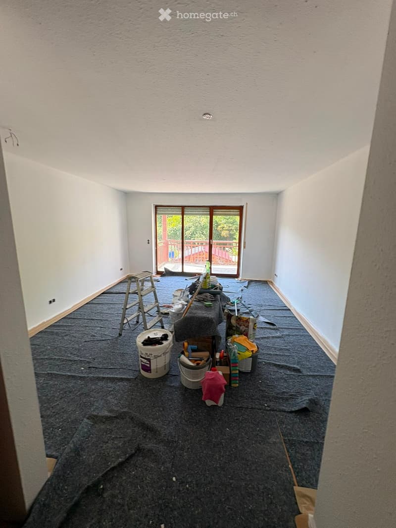 Empty room with white walls, carpeted floor, and a large window with a view of a deck and outdoor area. Construction materials and tools are scattered on the floor.
