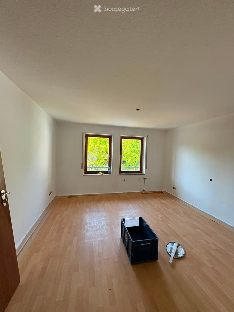 Empty room with wooden flooring, white walls, and two windows showing greenery outside. A black plastic crate and a small round tray with a paint roller are on the floor.