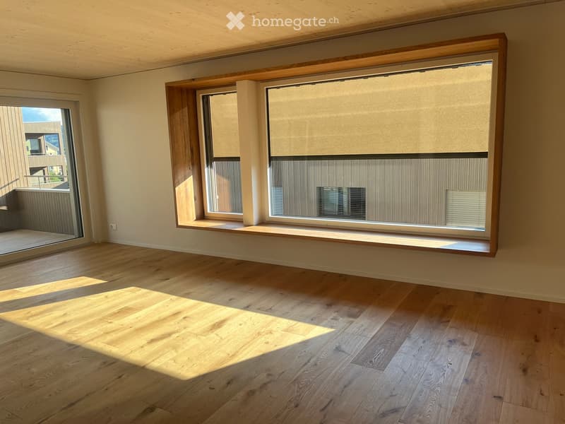 Empty room with wooden flooring, large windows with blinds, and a sliding glass door leading to a balcony.