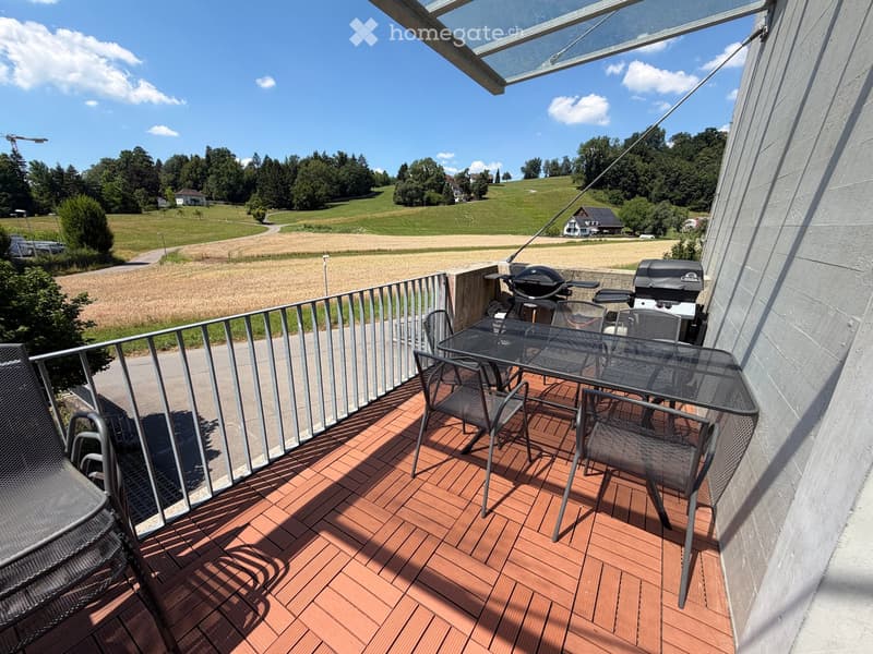 Balcony with wooden decking, metal railing, outdoor table and chairs, and two grills.