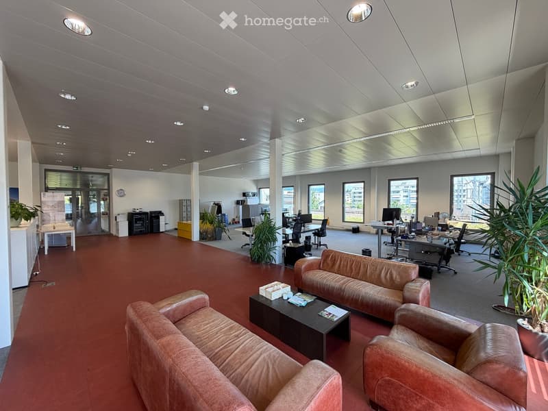 Modern office space with red carpeting, white walls, and a high ceiling. Features include a leather sofa, coffee table, office chairs, desks, and large windows with a view of the city.