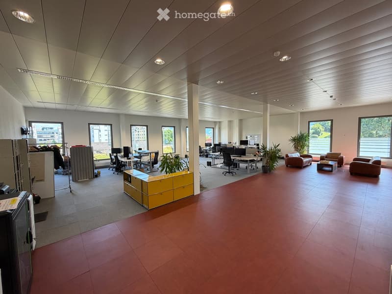 Modern office space with red tiled floor, large windows, desks, chairs, sofas, and plants.