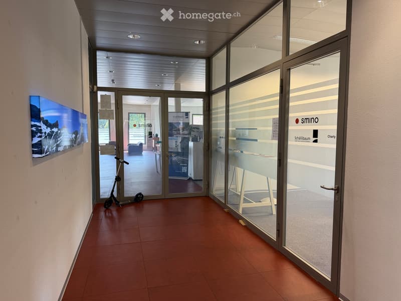 Corridor with glass doors leading to an office area, featuring a scooter parked near the entrance and a large wall-mounted display on the left side.