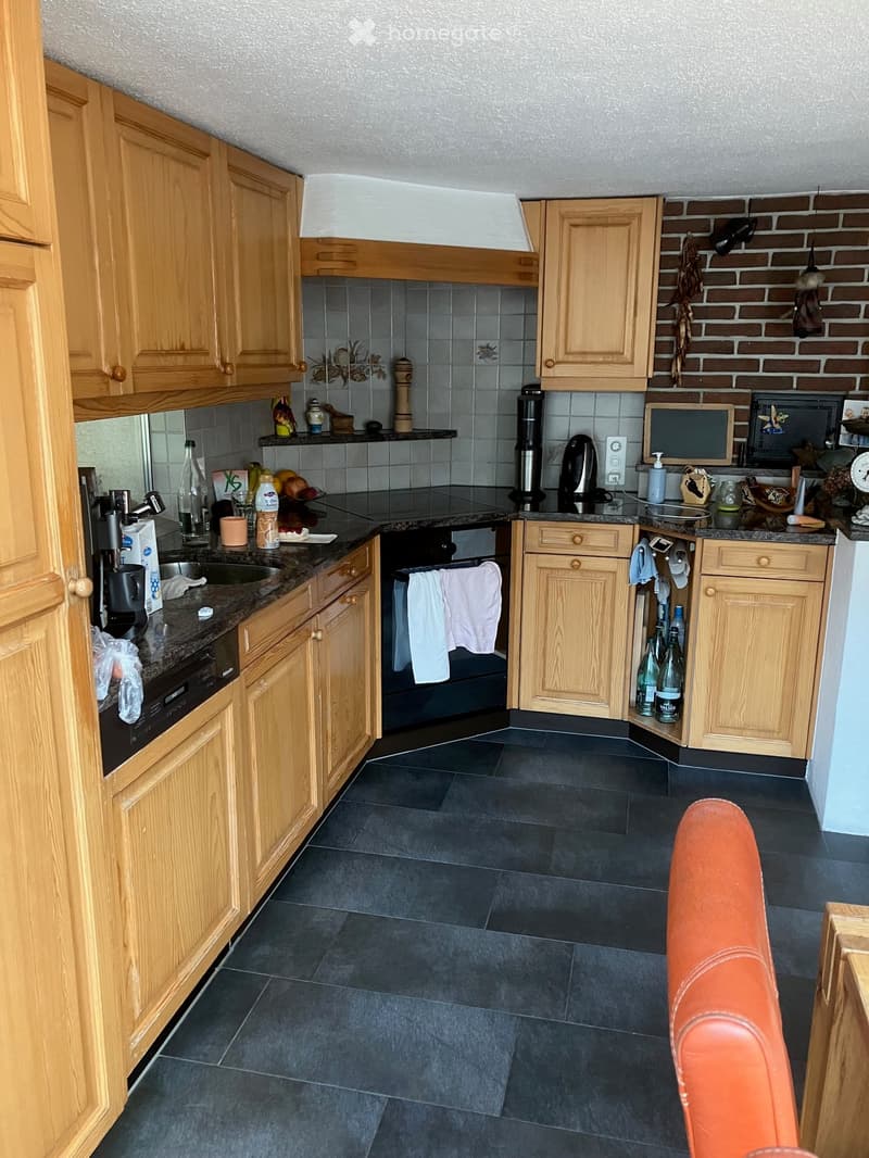 The kitchen features wooden cabinets, a dark tiled floor, and various appliances including a sink, oven, microwave, coffee maker, and fridge. The countertops hold items such as bottles, cups, and a microwave.