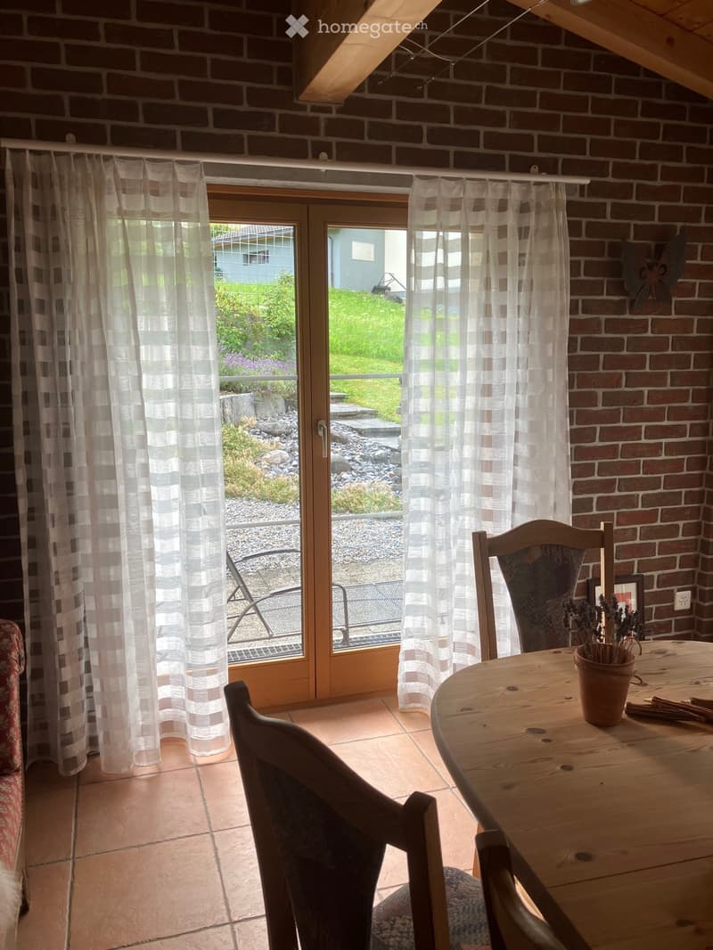 Bright kitchen with brick walls, large window with sheer curtains, wooden dining table with chairs, and a vase with dried flowers.