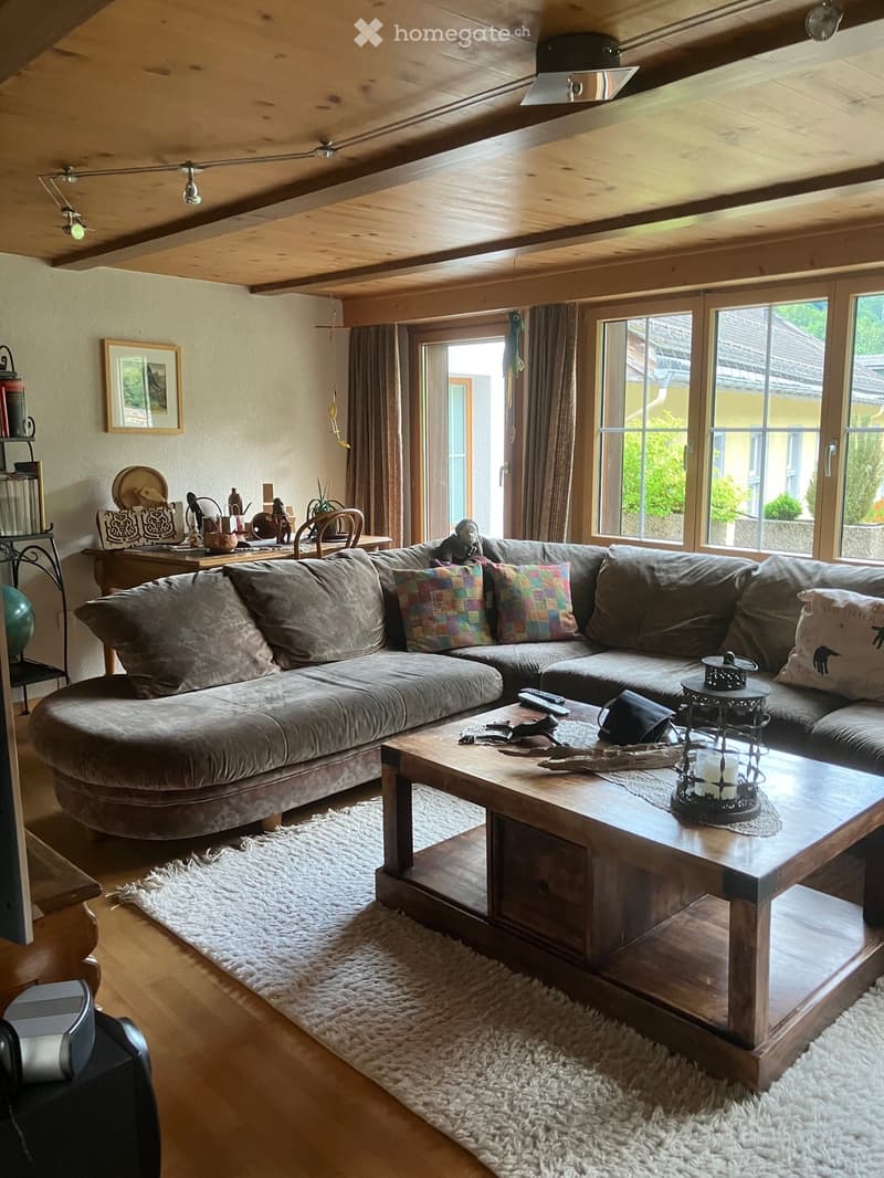 A cozy living room with a large sectional sofa, wooden coffee table, and warm lighting. The room features exposed wooden beams on the ceiling and large windows with curtains.