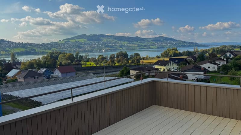 Balcony with wooden decking and metal railing, overlooking a scenic lake and town with surrounding hills.