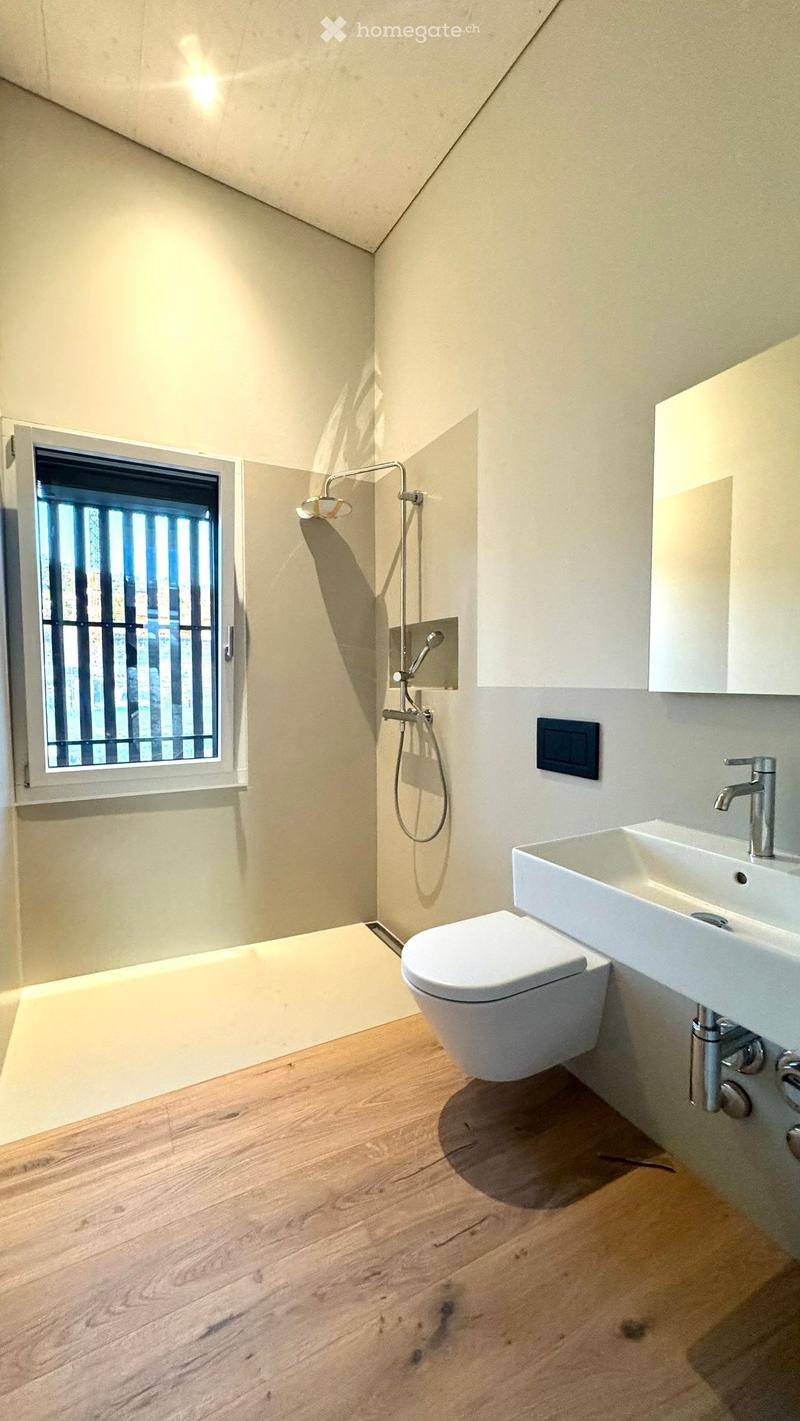 Modern bathroom with wooden flooring, white toilet, sink, and shower head. Features a window with vertical bars and a built-in cabinet.