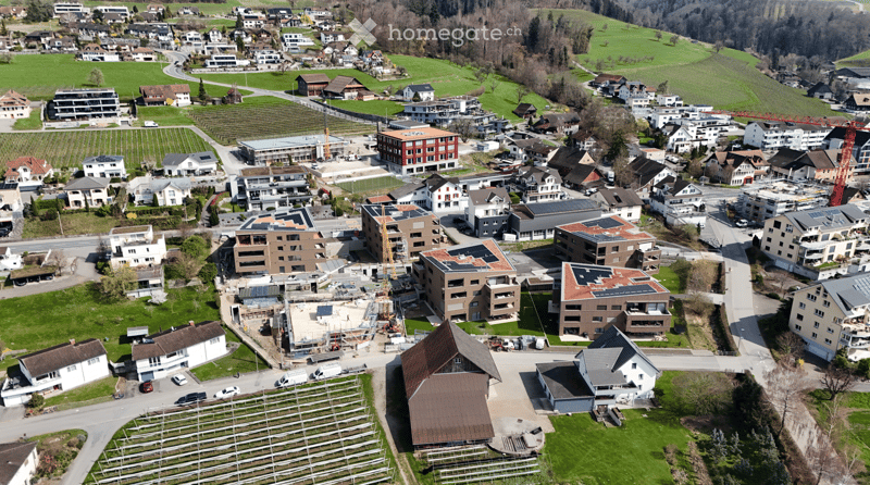 Aerial view of a residential area with multiple houses, apartment buildings, and green spaces. Some buildings have solar panels on their roofs, and there are construction cranes visible in the area.