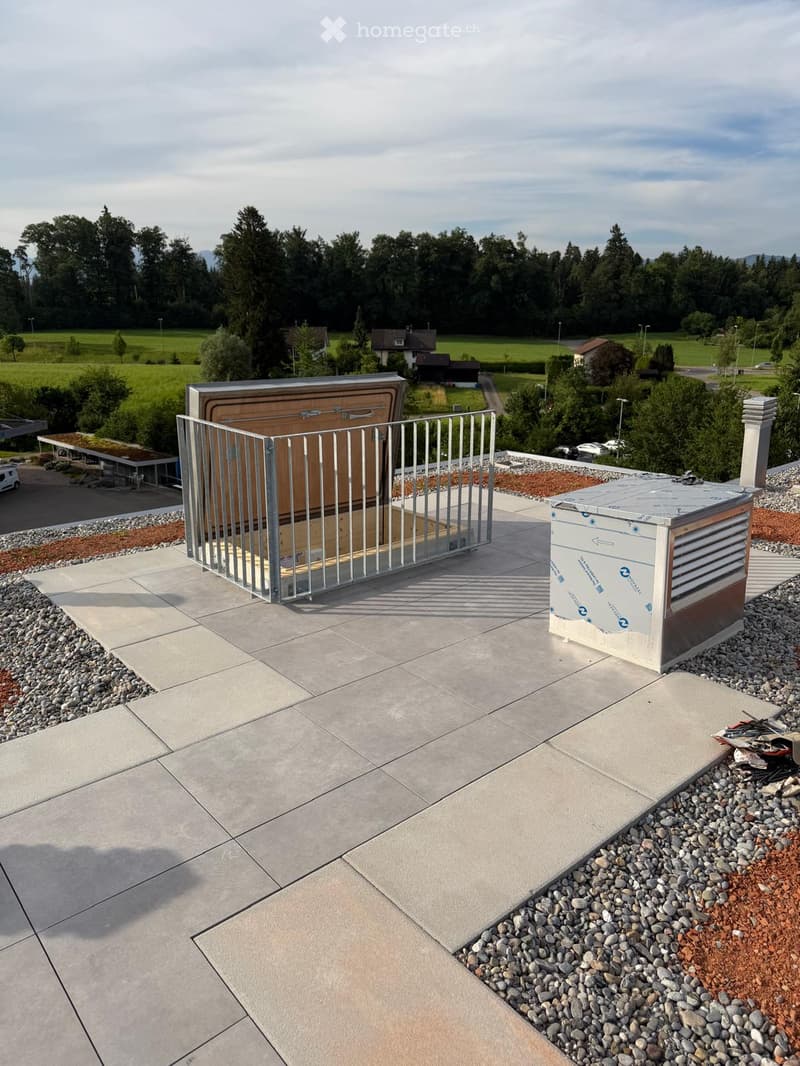 Roof terrace with concrete tiles, metal gate, and storage containers, surrounded by greenery and distant houses.