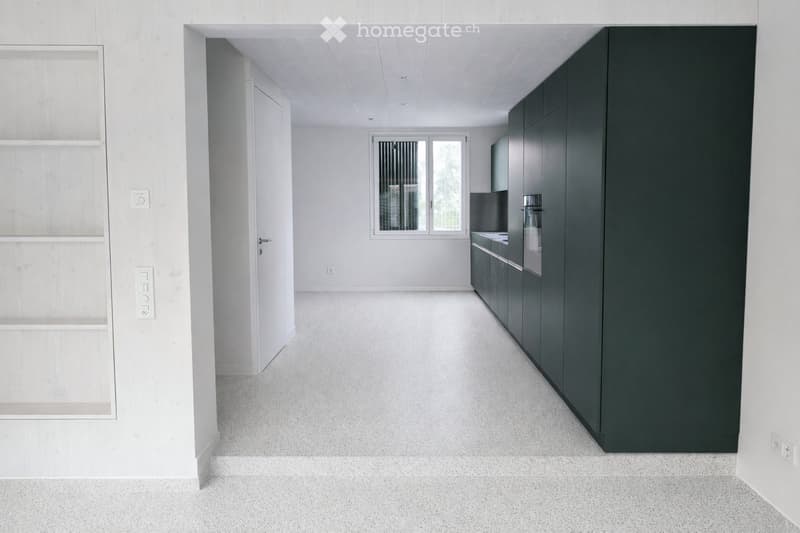 An empty, modern room with white walls, a grey carpeted floor, and a large window with blinds. The room features a dark-colored cabinet and a small sink area visible in the background.