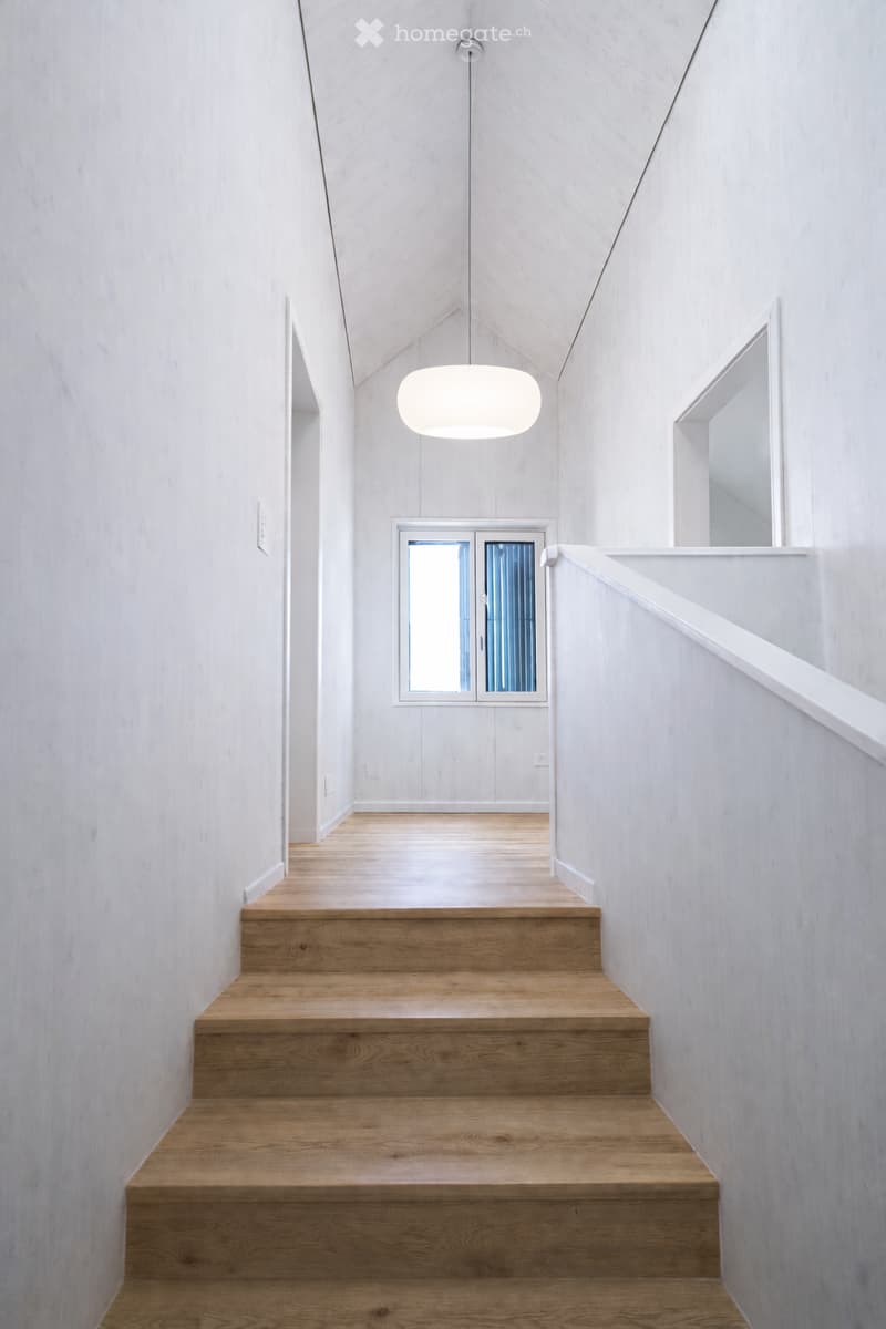 Modern, minimalist hallway with wooden stairs, white walls, and a pendant light fixture. The ceiling has a sloped design, and there is a window at the end of the hallway.
