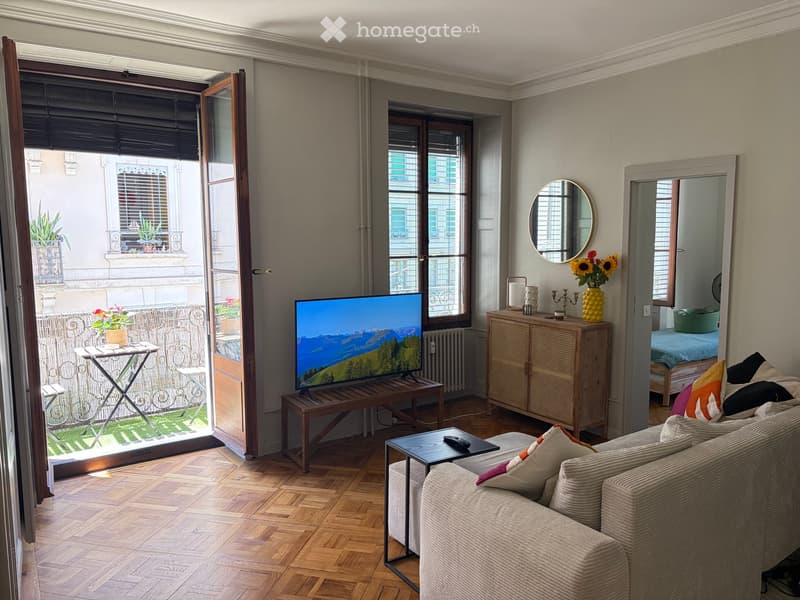 Living room with wooden flooring, large windows, balcony access, sofa, coffee table, television, side table, mirror, flower vase, and cabinet.