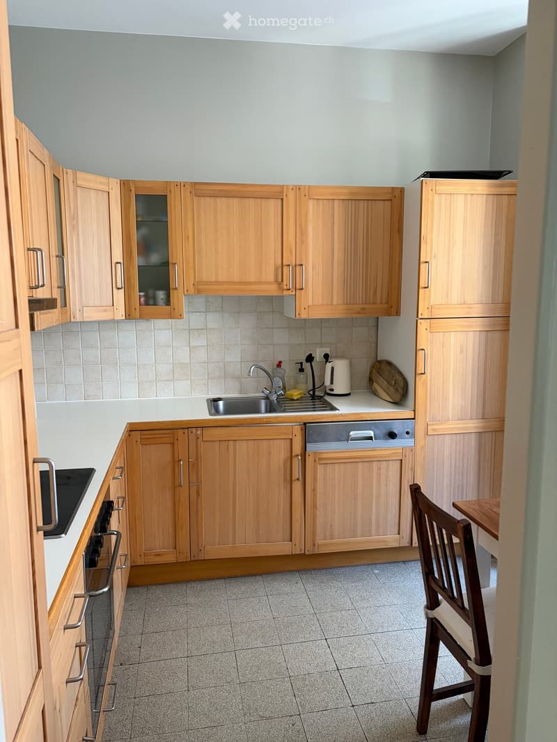 A modern kitchen with wooden cabinets, a sink, oven, dishwasher, refrigerator, and a small dining area with a chair and table.