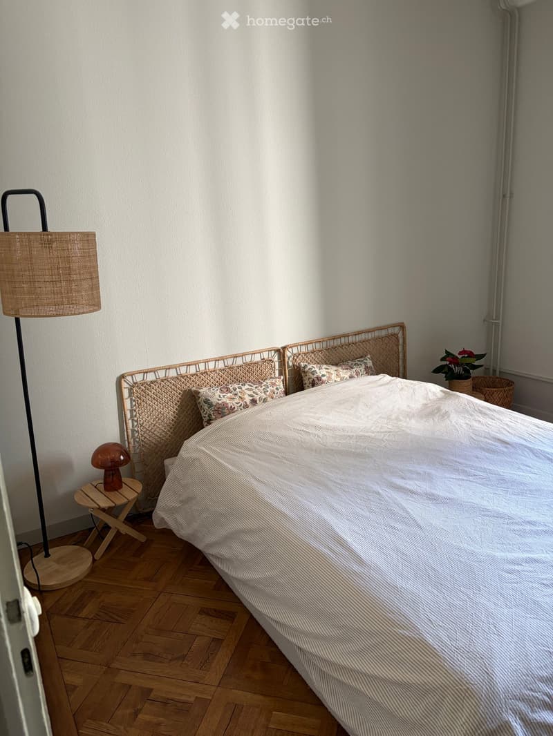 A cozy bedroom with a wooden floor, a wicker bed frame, a floor lamp, a small wooden stool with a table lamp, and a potted plant near the window.
