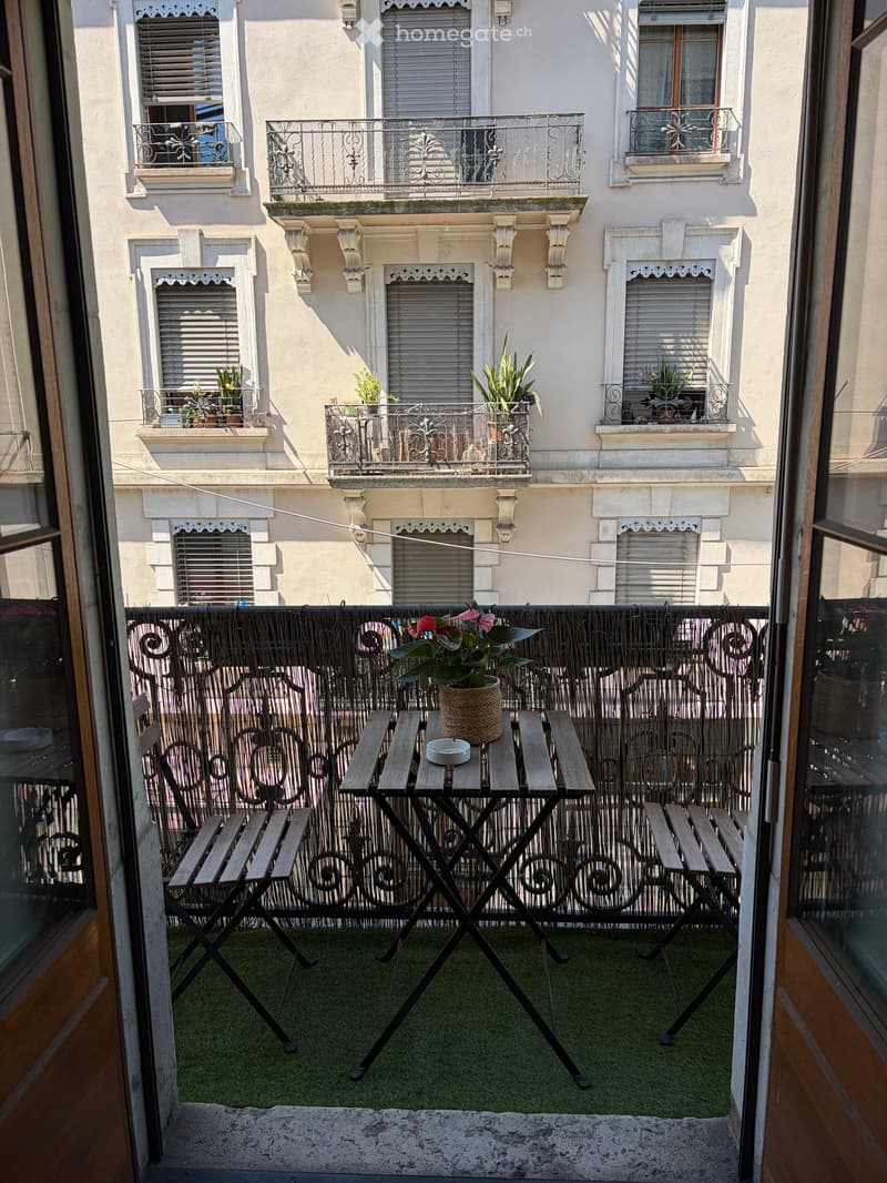 Balcony with a small table, two chairs, and a potted plant, overlooking a building with multiple balconies and windows.