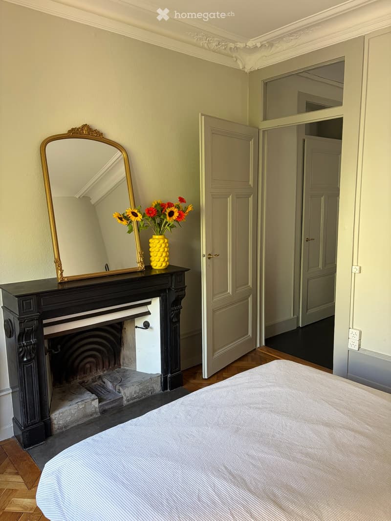 Elegant bedroom with a large bed, ornate black fireplace, decorative mirror, and a vase of sunflowers on the mantel.