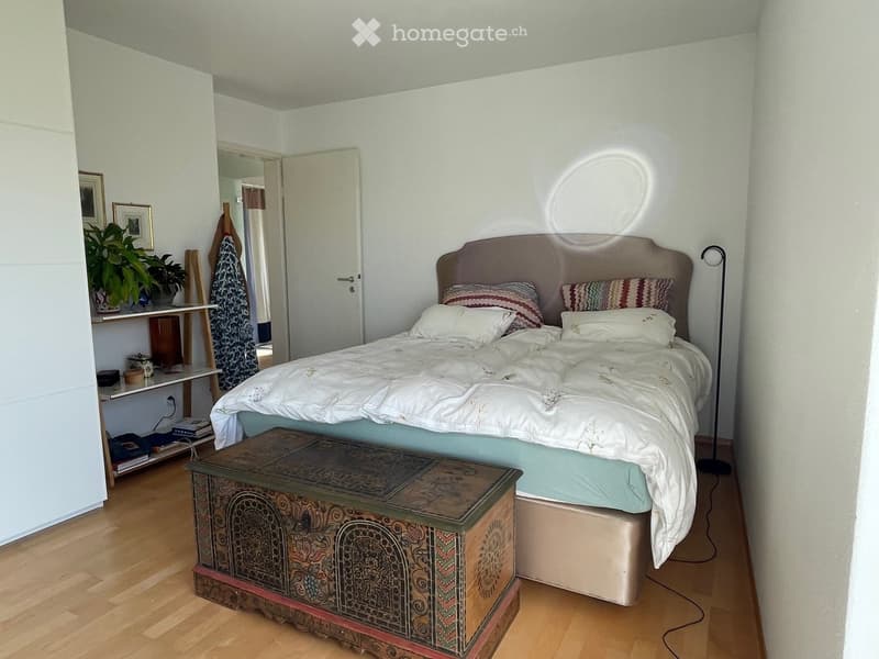 A well-lit bedroom with a large bed, patterned headboard, and decorative pillows. A wooden nightstand with a floor lamp is placed beside the bed, and a shelf with plants and picture frames is visible in the background.