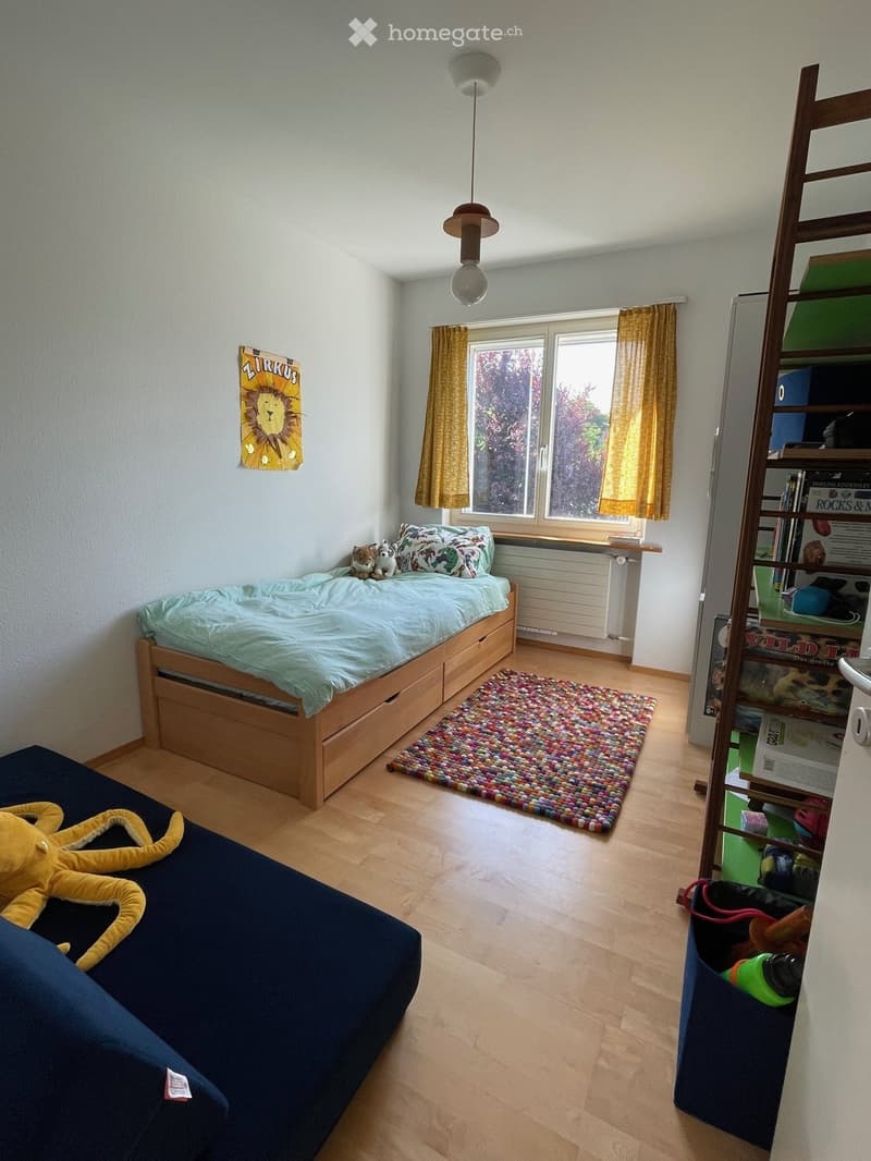 A small, well-lit bedroom with a single bed, colorful rug, and a shelf with books and toys. The room features wooden flooring and large windows with yellow curtains.