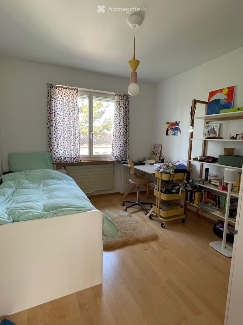 Cozy bedroom with wooden flooring, a bed with green bedding, a desk with a chair, a rolling cart, and a bookshelf filled with books and decorative items.