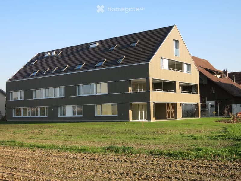 Modern two-story building with a sloped roof, large windows, and balconies, surrounded by a grassy lawn.