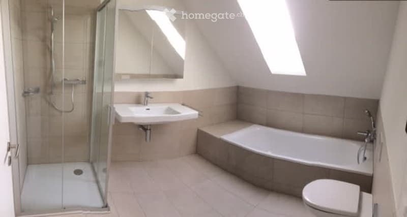 Modern bathroom with a bathtub, toilet, sink, mirror, and a glass-enclosed shower. The room features light-colored tiles and has skylight windows for natural light.