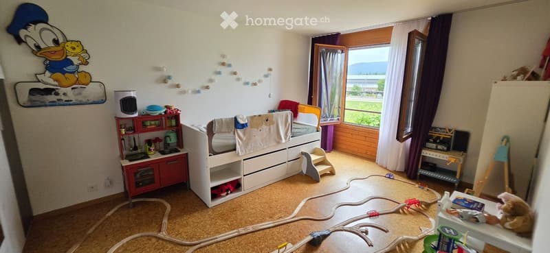 A child's bedroom with a white lofted bed, a toy train set on the floor, a small toy kitchen, a stool, a window with curtains, and various toys and decorations.