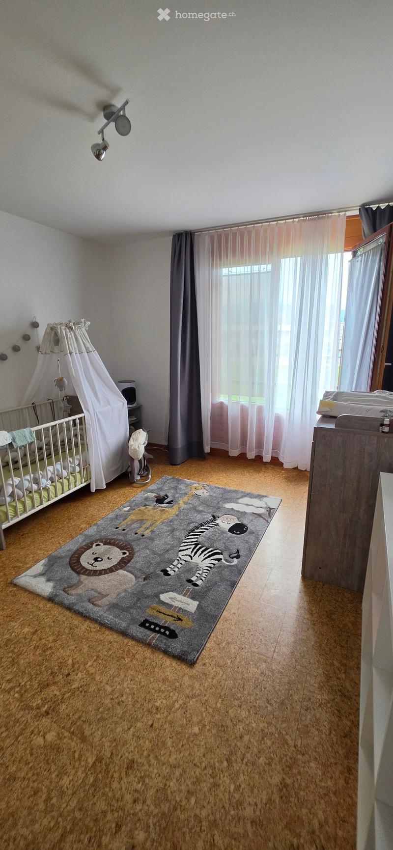 Brightly lit children's bedroom with wooden flooring, large window with curtains, crib, animal-themed rug, and wardrobe.