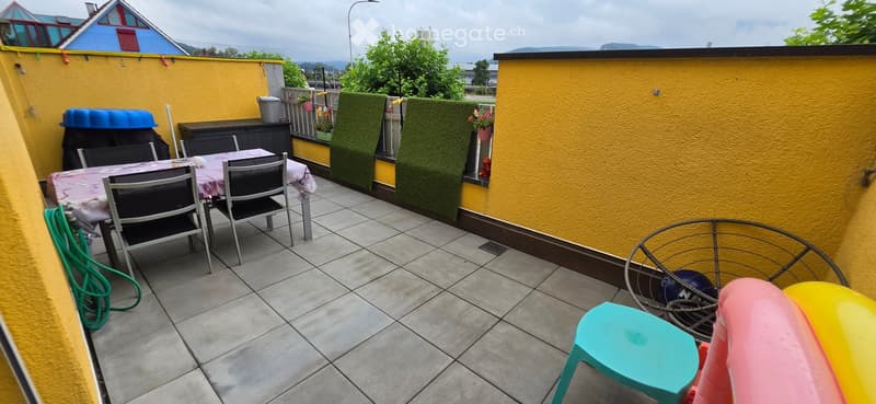 Balcony with tiled floor, yellow walls, dining table with chairs, green screen, barbecue, and colorful children's toys.