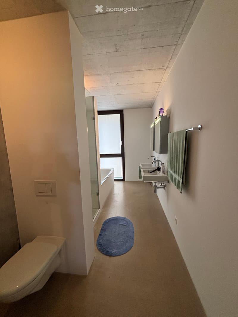 Modern bathroom with a toilet, sink, bathtub, and a towel on a rack. The space features a blue bath mat on the floor and a mirror above the sink.