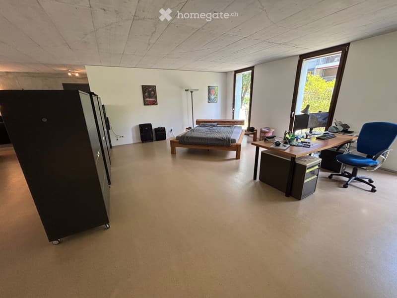 Open-plan room with a bed, office desk, office chair, computer, and a suitcase. Large windows allow natural light to fill the space.