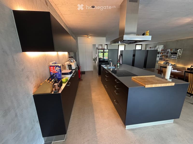 Modern kitchen with dark grey cabinets, a central island with an induction cooktop, and various appliances including a refrigerator, oven, and microwave. The countertop features a wooden cutting board and other small items.
