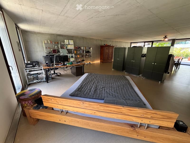 Open-plan room with a bed, desk, bookshelf, and cabinets. The space features a modern, minimalist design with concrete walls and large windows.
