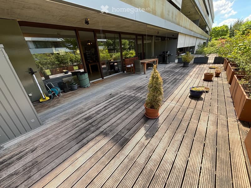 Wooden deck area with potted plants, outdoor furniture, and a view of the building interior through large glass doors.