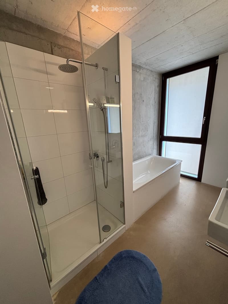 Modern bathroom with a glass-enclosed shower, bathtub, and sink. Features a large window providing natural light.