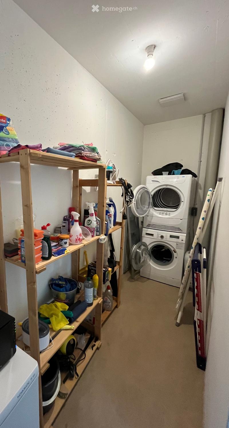 A narrow hallway with a wooden shelf on the left side holding various cleaning supplies and folded clothes, and a stacked washing machine and dryer on the right side.