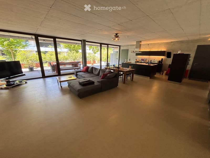 Open-plan living and dining area with large windows, grey sectional sofa, coffee table, dining table, and kitchen island.