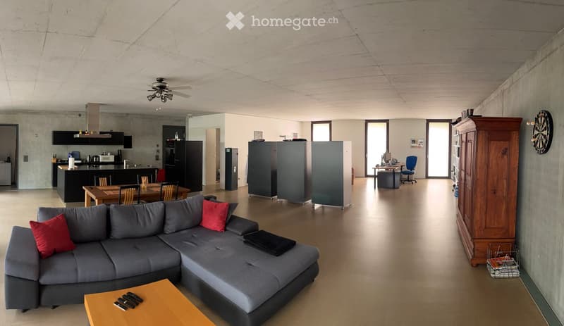 Open-plan living and dining area with a grey sectional sofa, coffee table, dining table with chairs, kitchen area with island, and office space with desk and chair. Features modern decor with red cushions and a dartboard on the wall.