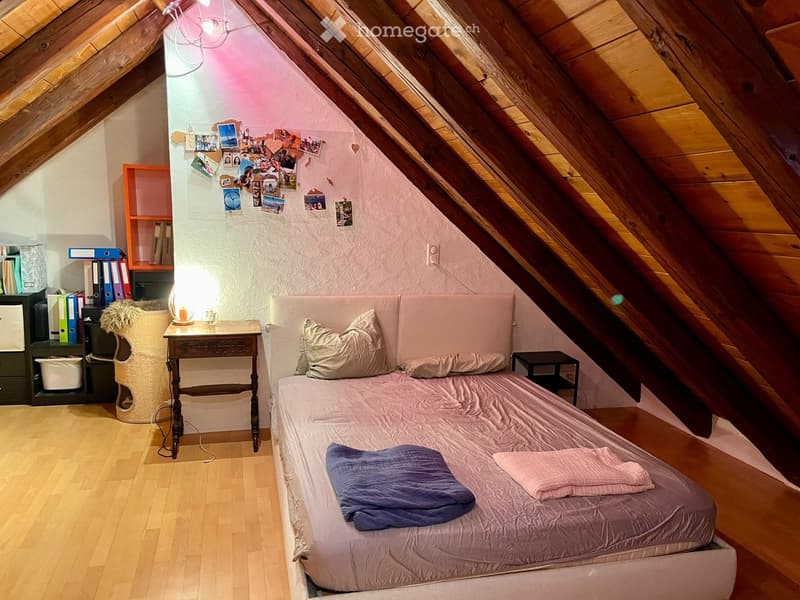 Cozy attic bedroom with wooden slanted ceilings, a single bed with pillows and blankets, a small wooden nightstand with a lamp, a wicker chair, and a shelf with books and decorative items.